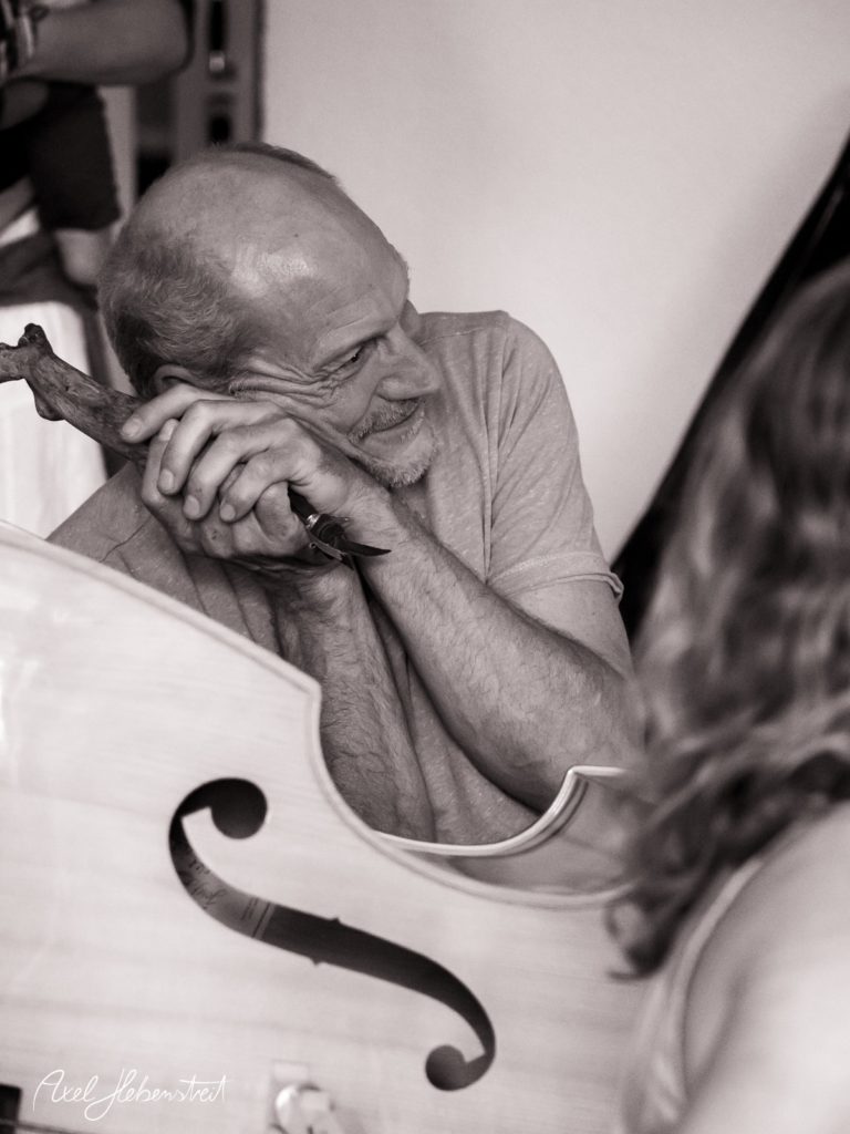 Ein musizierender Mann lehnt entspannt an einem Cello, während eine Person in der Nähe ihn anblickt – © Axel Hebenstreit