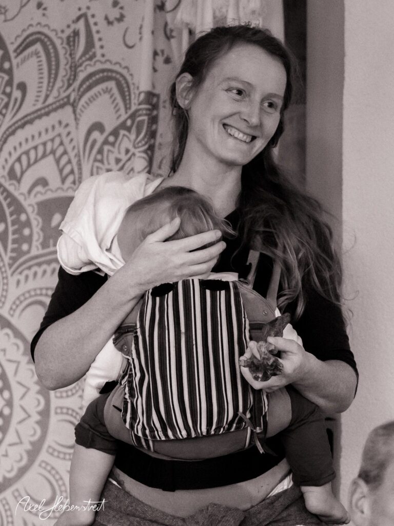 Eine lächelnde Mutter trägt ihr schlafendes Kind im Tragetuch und hält eine Blume in der Hand – © Axel Hebenstreit.