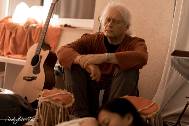 Ein meditierender Musiker umgeben von Instrumenten in einem harmonischen Raum – © Axel Hebenstreit.