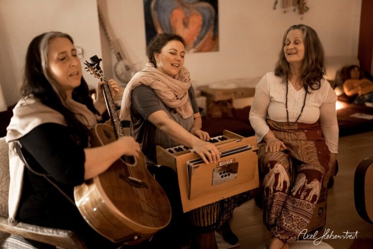 Drei Frauen musizieren gemeinsam, eine spielt Gitarre, eine andere ein Harmonium, in einer warmen und einladenden Umgebung. – © Axel Hebenstreit