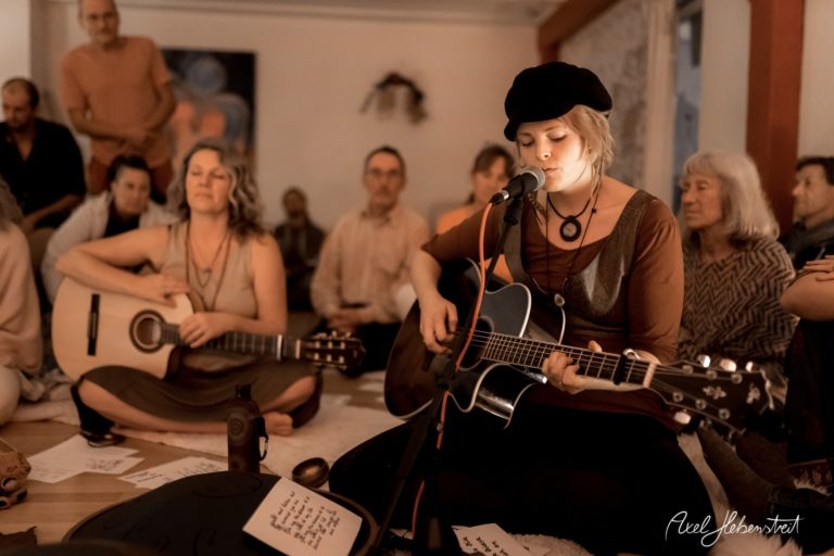 Frau mit Gitarre singt in einem vertrauten Kreis von Zuhörern, Atmosphäre von Gemeinschaft und Musik – © Axel Hebenstreit