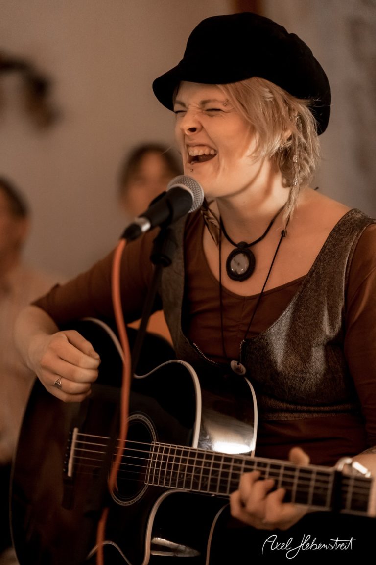 Eine Musikerin spielt mit Leidenschaft Gitarre und singt, während sie von einem Publikum umgeben ist – © Axel Hebenstreit.