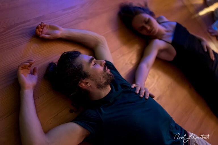 Zwei Personen liegen entspannt auf dem Boden und halten sich sanft an den Händen, in einer ruhigen und meditativen Atmosphäre. – © Axel Hebenstreit