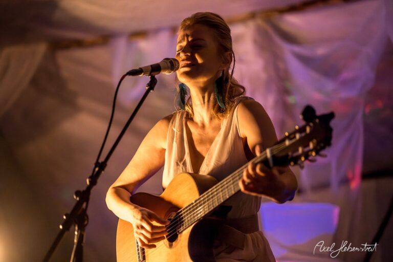 Eine Sängerin mit Gitarre performt leidenschaftlich auf einer Bühne in einem stimmungsvollen, beleuchteten Raum – © Axel Hebenstreit