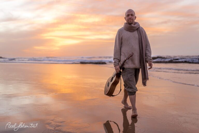 Mann mit Gitarre läuft barfuß am Strand bei Sonnenuntergang entlang – © Axel Hebenstreit