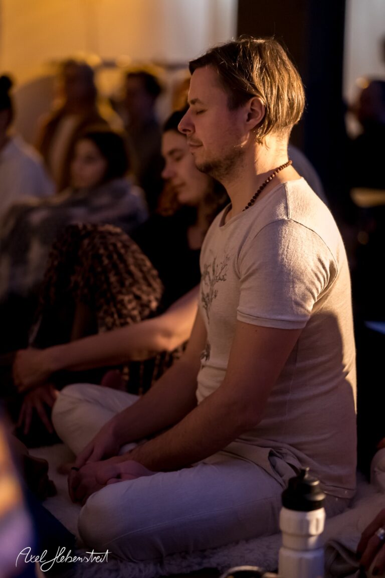 Eine meditative Person sitzt im Schneidersitz, umgeben von einer entspannten Atmosphäre bei einer Yoga-Veranstaltung – © Axel Hebenstreit