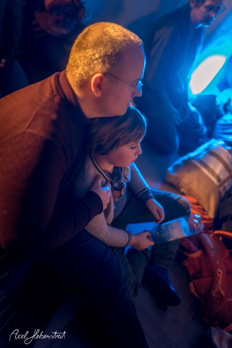 Ein erwachsener Mann umarmt einen Jungen in einer entspannten, blauen Lichtstimmung. – © Axel Hebenstreit