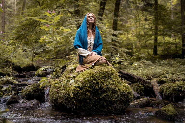 Frau in blauem Tuch sitzt meditativ auf moosbedecktem Stein in idyllischem Waldbach – © Axel Hebenstreit