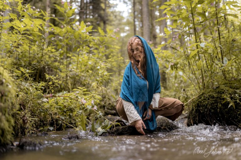 Frau mit blauem Tuch kniet am Bach im grünen Wald und blickt nachdenklich zur Seite – © Axel Hebenstreit
