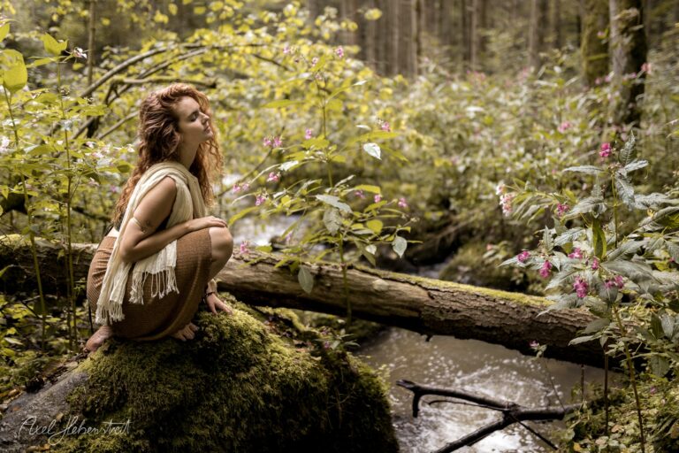 Nachdenkliche Frau in Naturkleidung sitzt auf moosigem Baumstamm an waldreichem Bach mit Wildblüten – © Axel Hebenstreit