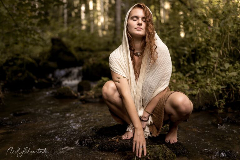 Junge Frau mit langen roten Haaren in weißem Tuch hockt nachdenklich an einem Waldbach – © Axel Hebenstreit