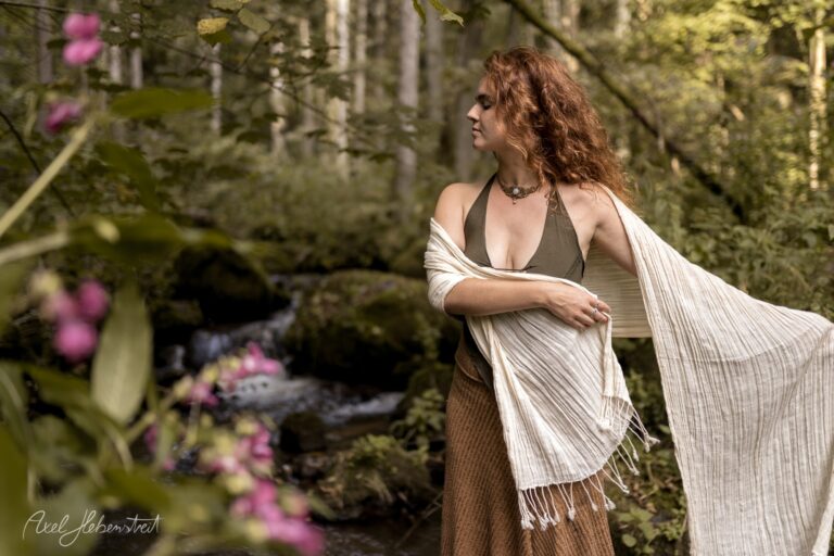 Rothaarige Frau in weißem Tuch und braunem Kleid posiert anmutig in einem moosigen Waldgang mit rosa Blüten – © Axel Hebenstreit