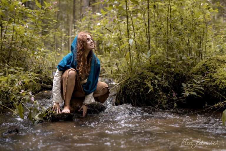 Junge Frau mit roten Locken in blauem Umhang hockt nachdenklich in einem Waldbach umgeben von grüner Vegetation – © Axel Hebenstreit