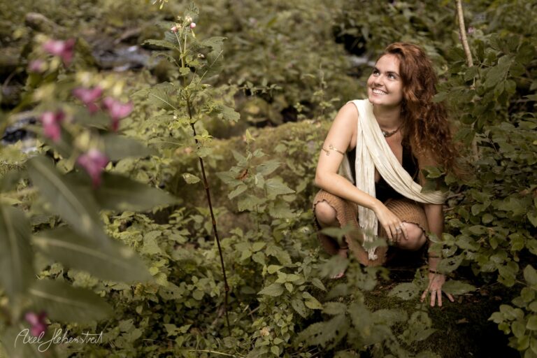Lächelnde Frau hockt inmitten von grünen Pflanzen und rosa Blüten in naturbelassener Gartenumgebung – © Axel Hebenstreit