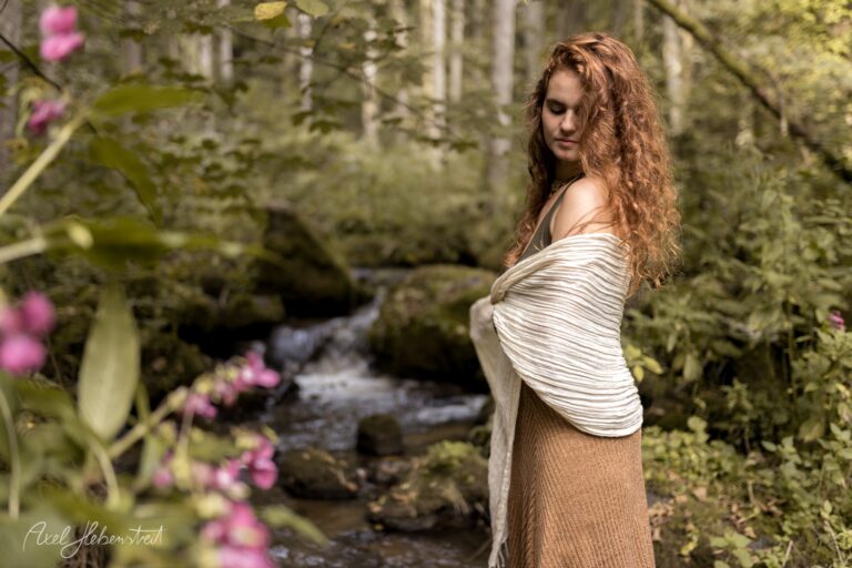 Junge Frau mit lockigem rötlichem Haar in cremefarbenem Tuch posiert an waldreichem Bachbett mit pinken Blüten – © Axel Hebenstreit