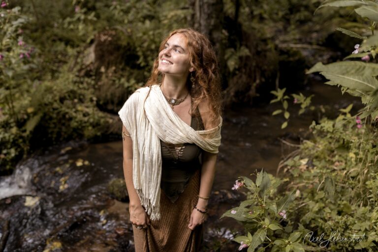 Junge Frau in mittelalterlicher Tracht blickt selig nach oben, umgeben von Wildblumen an einem Waldbach – © Axel Hebenstreit