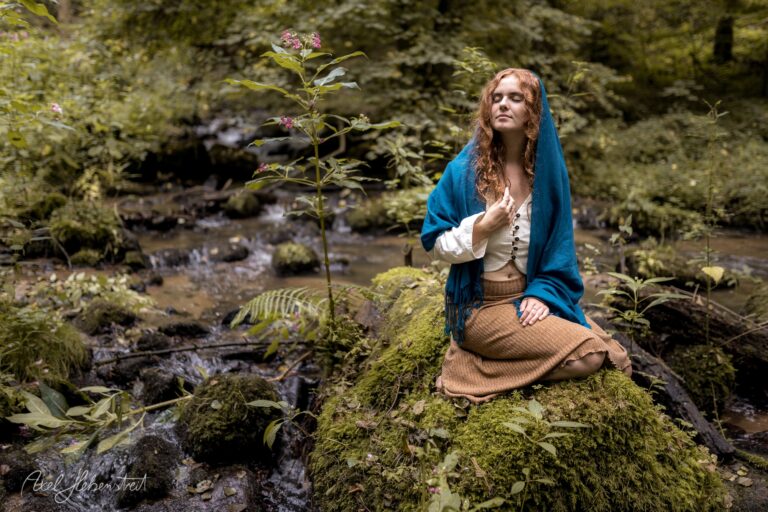 Frau mit rötlichem Haar in blauem Umhang sitzt meditativ an einem moosigen Bach im Wald – © Axel Hebenstreit