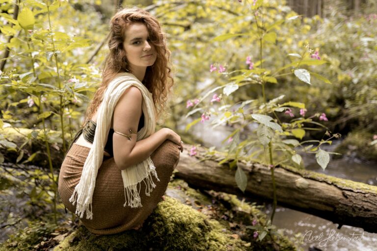 Junge Frau mit lockigem Haar in weißem Tuch sitzt in blühendem Waldbach umgeben von Natur – © Axel Hebenstreit