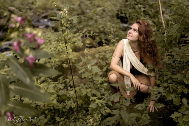 Frau in weißem Tuch hockt nachdenklich in üppigem Garten mit rosa Blüten – © Axel Hebenstreit