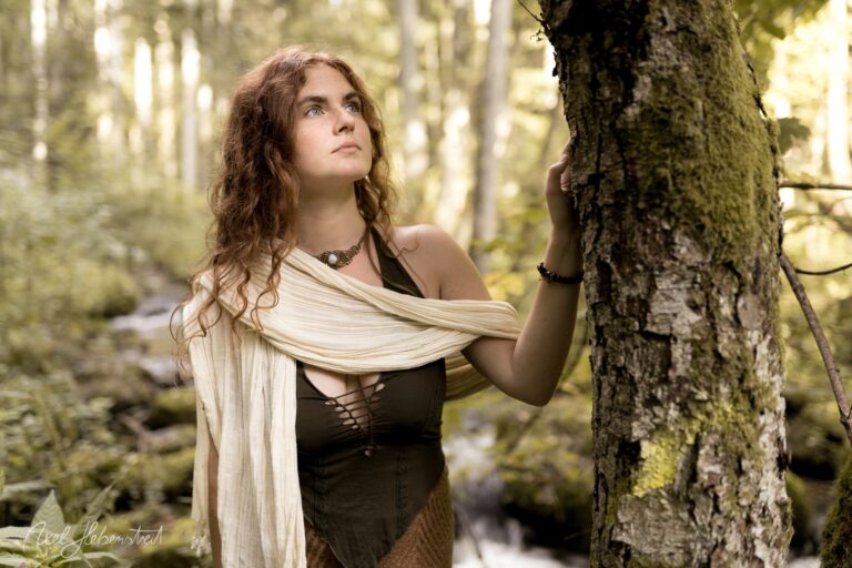 Junge Frau mit lockigem Haar in mittelalterlicher Kleidung lehnt an moosigem Baum im Wald – © Axel Hebenstreit