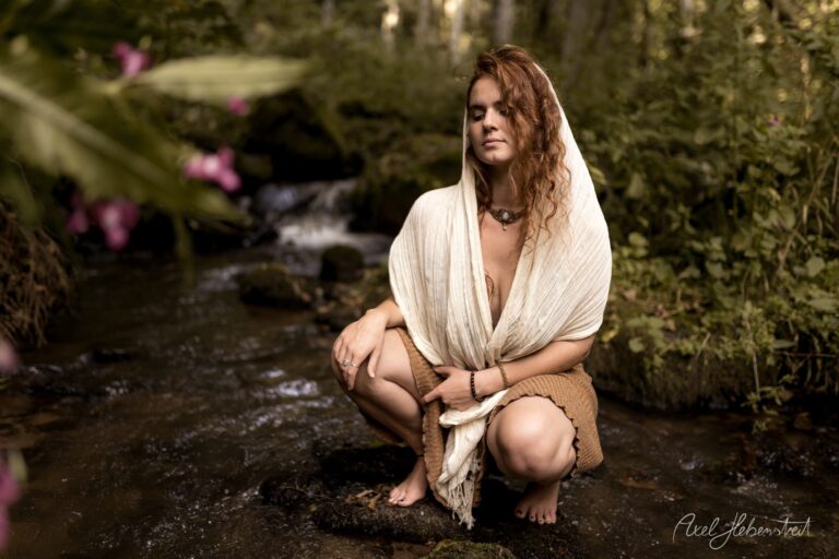 Frau in weißem Tuch sitzt meditativ an einem Bach in grüner Naturlandschaft mit rosa Blüten – © Axel Hebenstreit