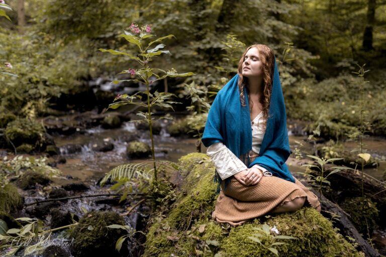 Junge Frau mit rötlichen Haaren und blauem Tuch sitzt nachdenklich auf moosigem Stein an einem Bach im Wald – © Axel Hebenstreit