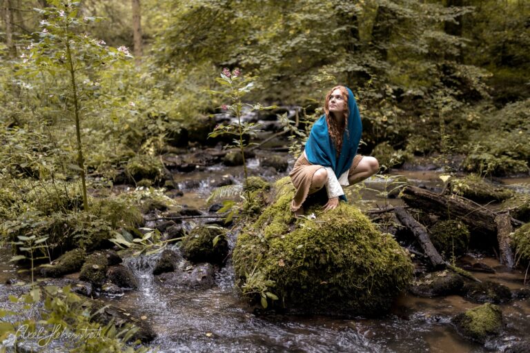 Frau mit blauem Kopftuch sitzt nachdenklich auf moosbedecktem Stein an einem Waldbach – © Axel Hebenstreit