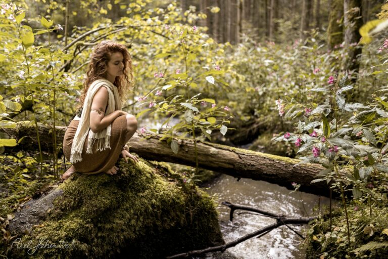 Frau mit lockigem Haar sitzt nachdenklich auf moosigem Stein neben Waldbach, umgeben von blühenden Wildkräutern – © Axel Hebenstreit
