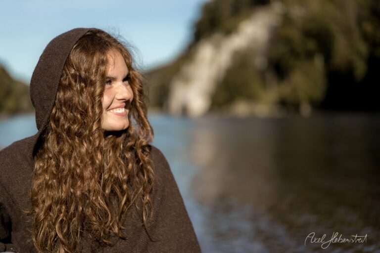 Porträt einer lächelnden Frau mit langen lockigen braunen Haaren in brauner Kapuzenjacke an einem See mit Felsklippen im Hintergrund – © Axel Hebenstreit