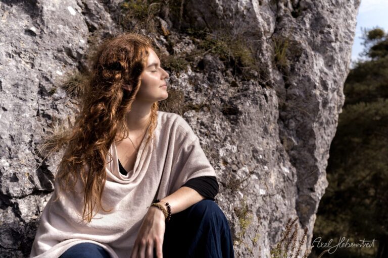 Junge Frau mit langen braunen Haaren sitzt nachdenklich an einer alten Steinmauer – © Axel Hebenstreit