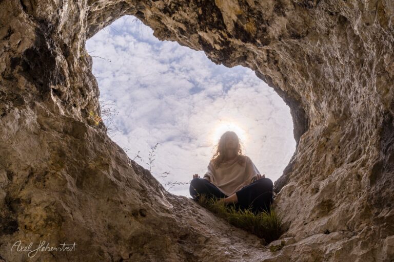 Person sitzt meditativ in einer Felshöhle und blickt zum bewölkten Himmel – © Axel Hebenstreit