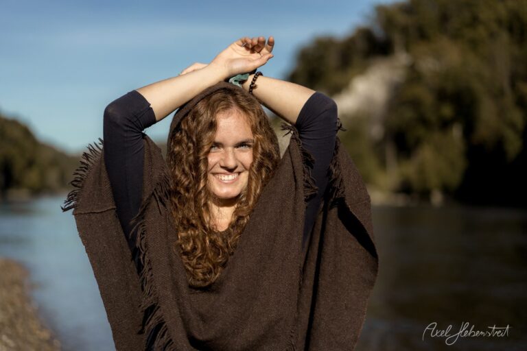 Junge Frau mit lockigem braunem Haar lächelt entspannt an einem Fluss mit Fransen-Poncho – © Axel Hebenstreit