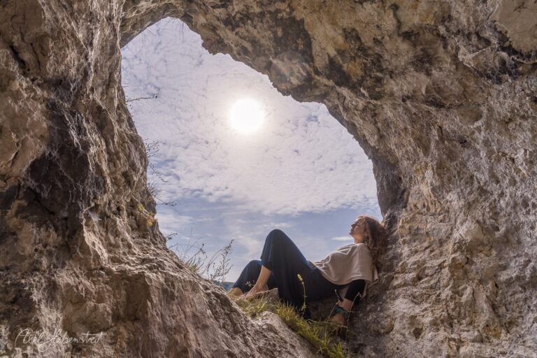 Frau sitzt in einer Felshöhle und blickt auf bewölkten Himmel und Landschaft – © Axel Hebenstreit
