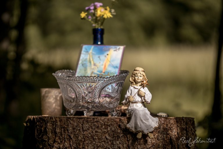 Engelsfigur sitzt auf Baumstumpf neben ornamentaler Vase mit Blumen und Kunstwerk in natürlicher Umgebung – © Axel Hebenstreit