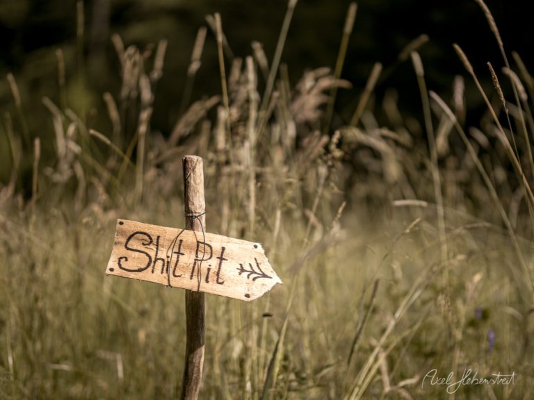 Holzschild mit der Aufschrift Shit Pit zeigt humorvoll in ein verwildertes Grasfeld – © Axel Hebenstreit