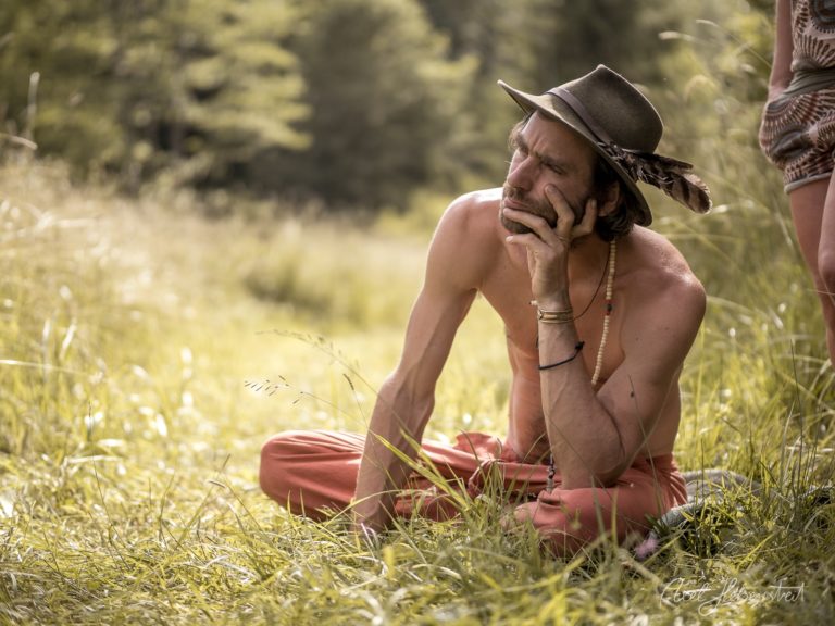 Mann mit Cowboyhut sitzt nachdenklich in grüner Wiese und blickt in die Ferne – © Axel Hebenstreit