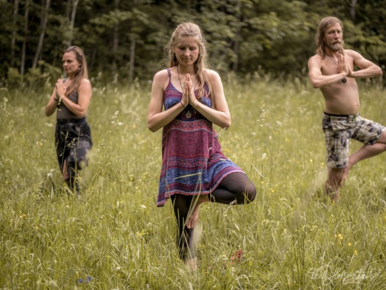 Drei Personen praktizieren Yoga-Positionen in einer blütenreichen Wiese vor Waldkulisse – © Axel Hebenstreit