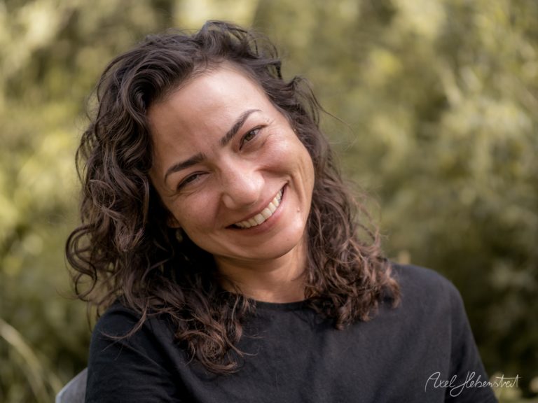 Porträt einer lächelnden Frau mit lockigem braunem Haar vor unscharfem grünem Naturhintergrund – © Axel Hebenstreit