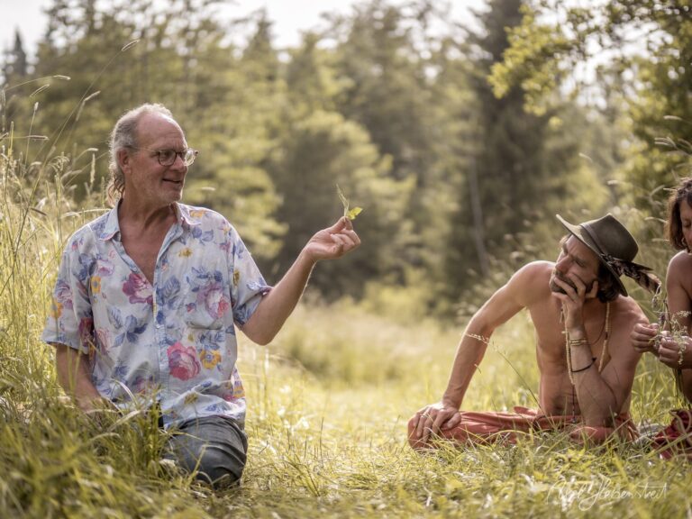 Ein älterer Mann mit Brille zeigt einer sitzenden Person in freier Natur Wildkräuter, umgeben von grünen Wiesen und Wald – © Axel Hebenstreit