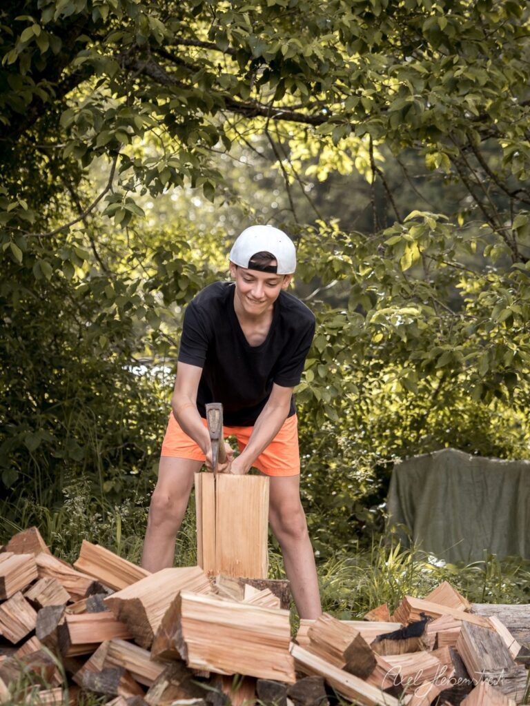 Junger Mann spaltet Holz mit Axt in grüner Natur unter Baumkrone – © Axel Hebenstreit
