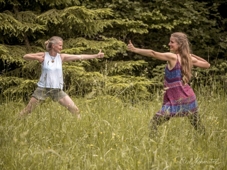 Zwei Frauen in Yoga-Pose in einer blütenreichen Wiese vor Nadelwald in freudiger Verbundenheit – © Axel Hebenstreit