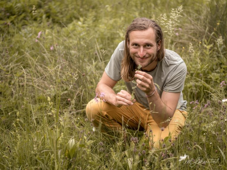 Mann mit langen Haaren kniet in blühendem Wildkrautfeld und lächelt in die Kamera – © Axel Hebenstreit