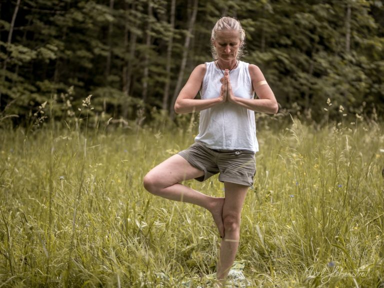 Frau in weißem Yoga-Top praktiziert Tree Pose in einer Bergwiese mit Wald im Hintergrund – © Axel Hebenstreit