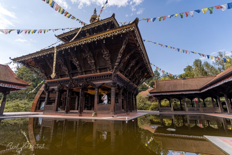 Ornamentale buddhistische Tempelanlage mit aufwendig verziertem Hauptgebäude spiegelt sich im Wasser wider, bunte Gebetsfahnen flattern im Wind – © Axel Hebenstreit