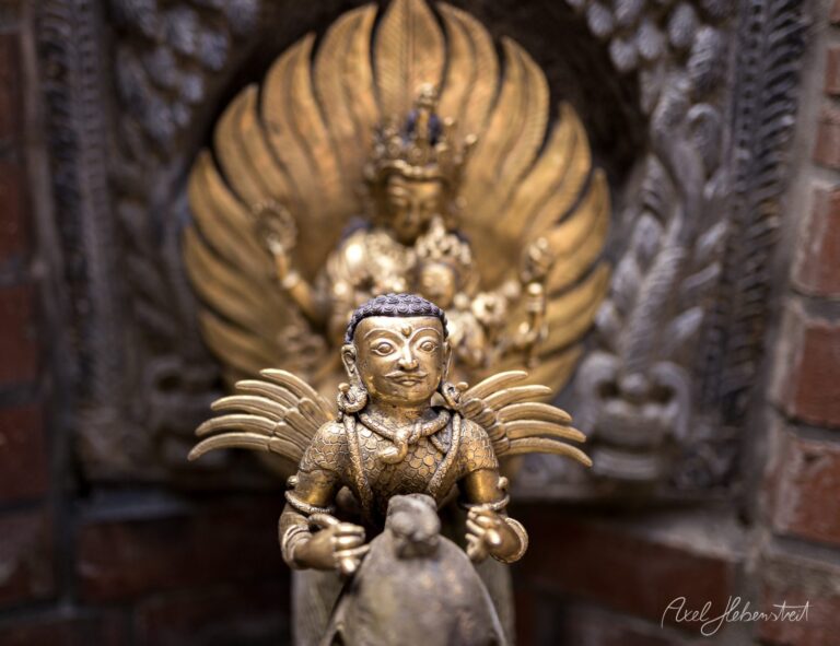 Vergoldete buddhistische Statue mit Flügeln im Fokus vor großem Hintergrund-Relief in warmen Goldtönen – © Axel Hebenstreit