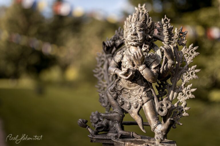 Detaillierte Bronzestatue einer mehrarmeigen göttlichen Figur mit ornamentalem Flammenkranz vor unscharfem Garten – © Axel Hebenstreit