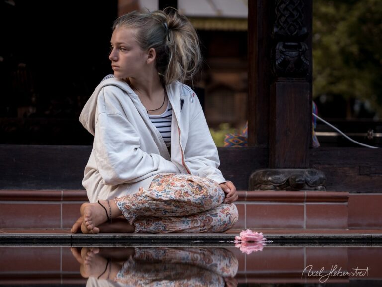 Nachdenkliches Mädchen sitzt in meditativer Pose an einem Gewässer in einem asiatischen Tempel mit Blüte im Wasser – © Axel Hebenstreit