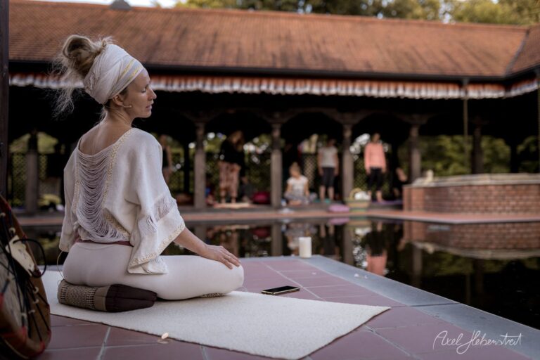 Frau in Yoga-Pose sitzt meditativ auf einer Matte vor einem historischen Gebäude mit Wasserbecken – © Axel Hebenstreit