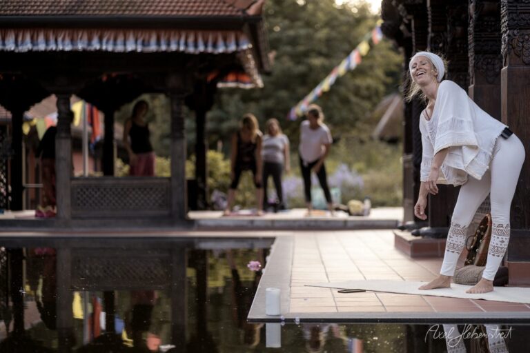 Lächelnde Frau in weißem Yoga-Outfit in entspannter Pose an einem asiatischen Tempel mit Wasserspiegelung – © Axel Hebenstreit