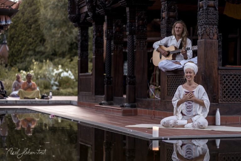 Zwei Frauen in weißer Kleidung meditieren und musizieren an einem See vor asiatischer Architektur in friedvoller Atmosphäre – © Axel Hebenstreit